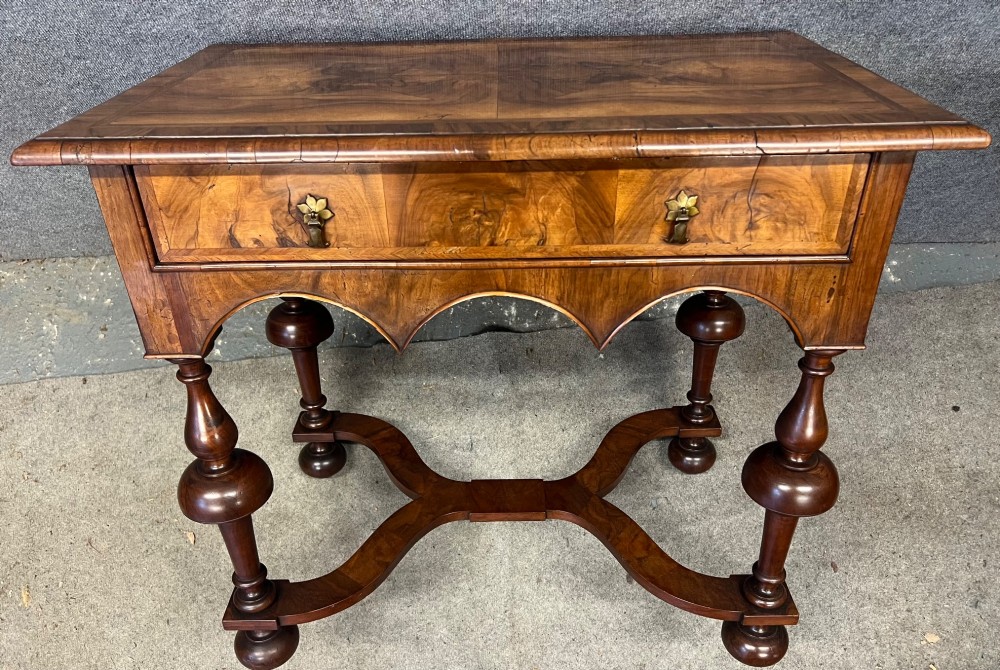 A William and Mary style walnut lowboy table featuring a quartered figured veneer top, a single drawer with brass drop handles, a shaped apron, turned legs, and a serpentine stretcher. View III