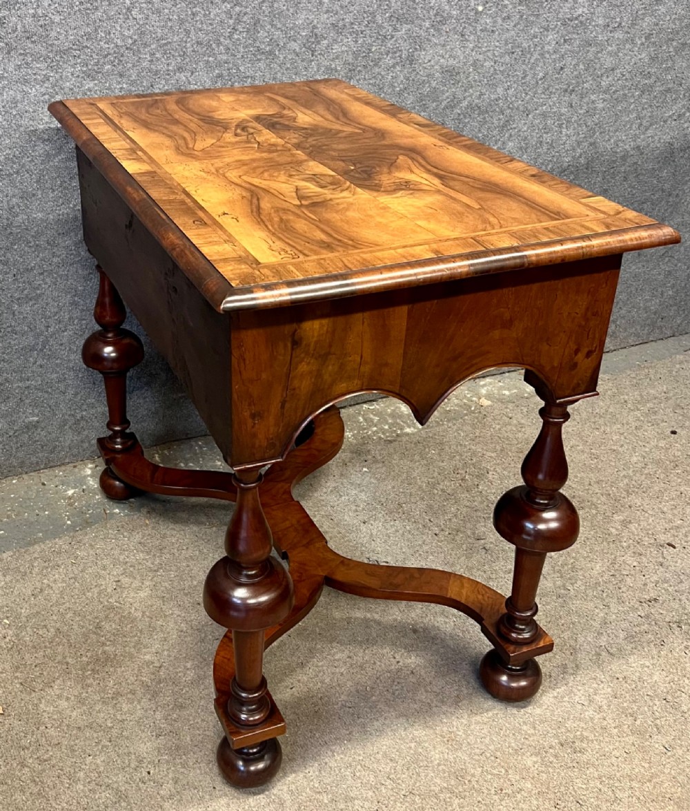A William and Mary style walnut lowboy table featuring a quartered figured veneer top, a single drawer with brass drop handles, a shaped apron, turned legs, and a serpentine stretcher. View V