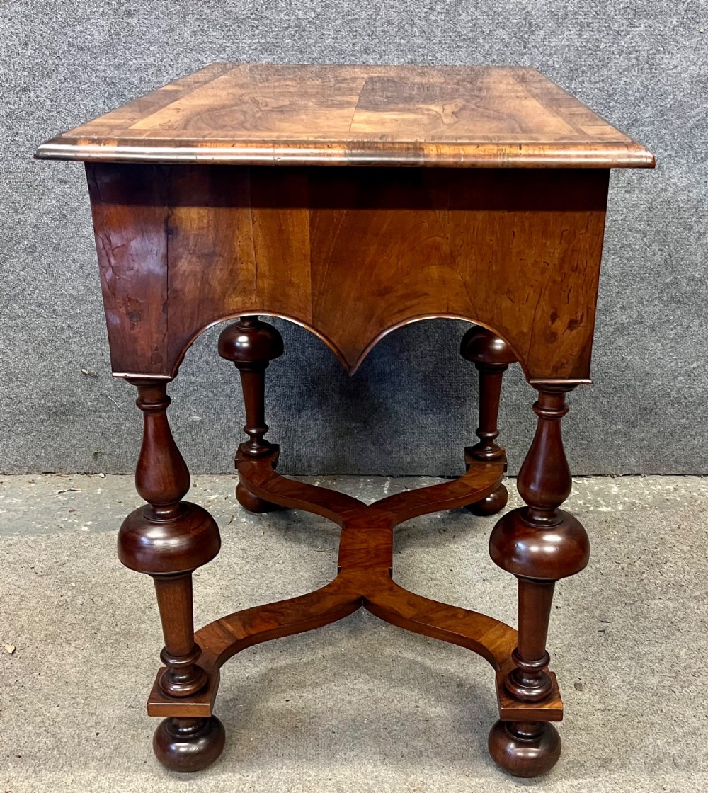 A William and Mary style walnut lowboy table featuring a quartered figured veneer top, a single drawer with brass drop handles, a shaped apron, turned legs, and a serpentine stretcher. View VI