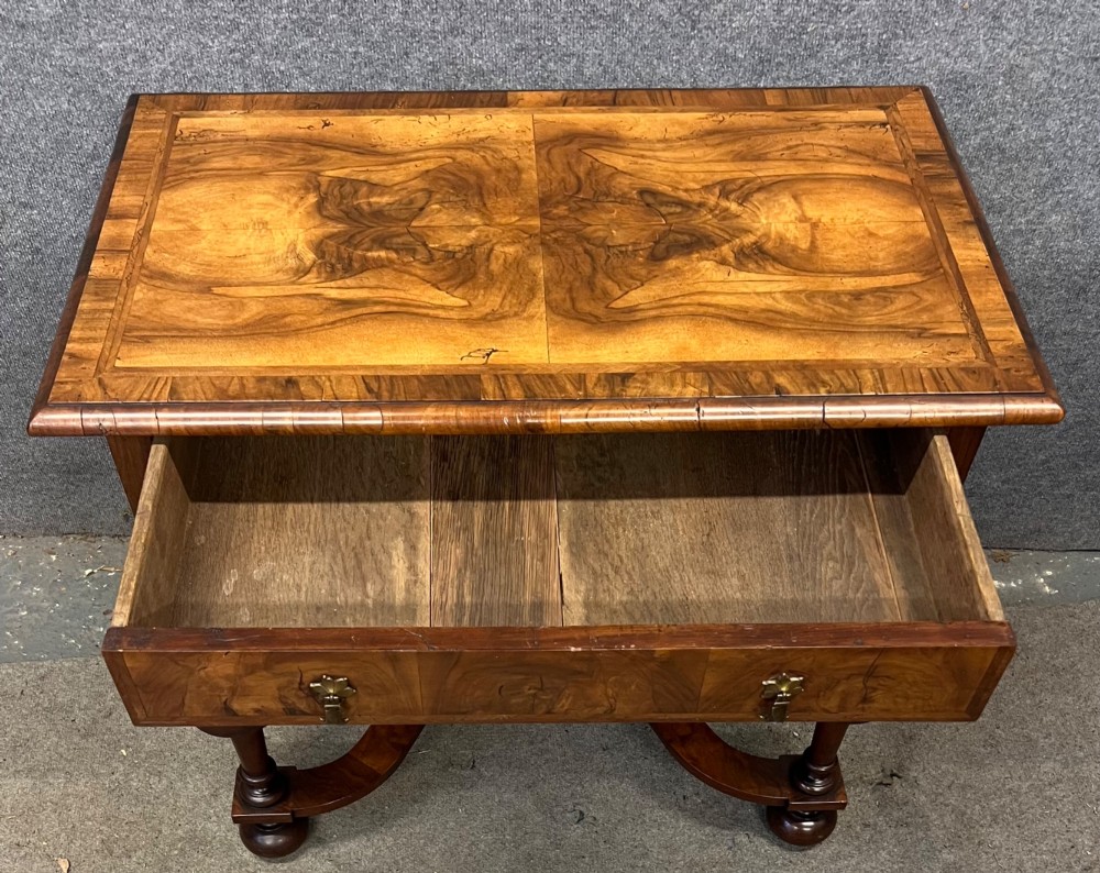 A William and Mary style walnut lowboy table featuring a quartered figured veneer top, a single drawer with brass drop handles, a shaped apron, turned legs, and a serpentine stretcher. View X