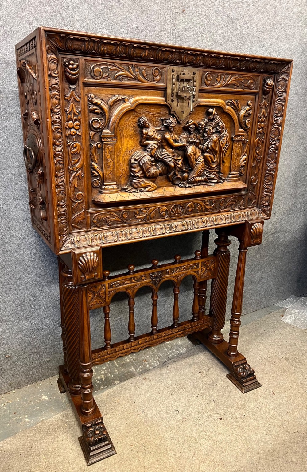 A carved Spanish vargueno cabinet in dark wood, featuring relief figures on the drop-front flap, decorative brass hardware, and a turned trestle stand.