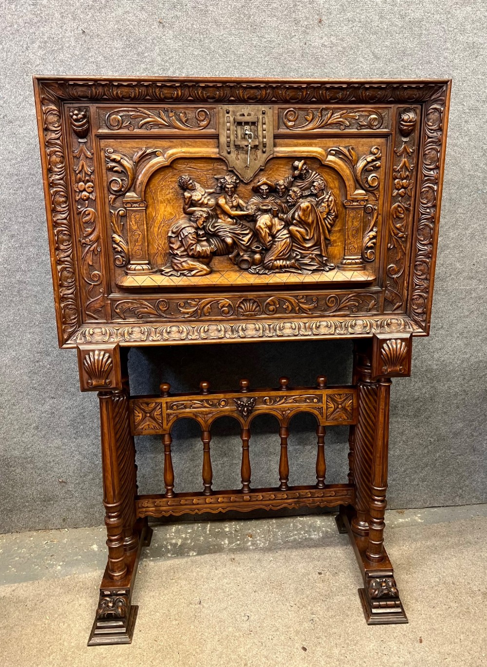 A carved Spanish vargueno cabinet in dark wood, featuring relief figures on the drop-front flap, decorative brass hardware, and a turned trestle stand. View II