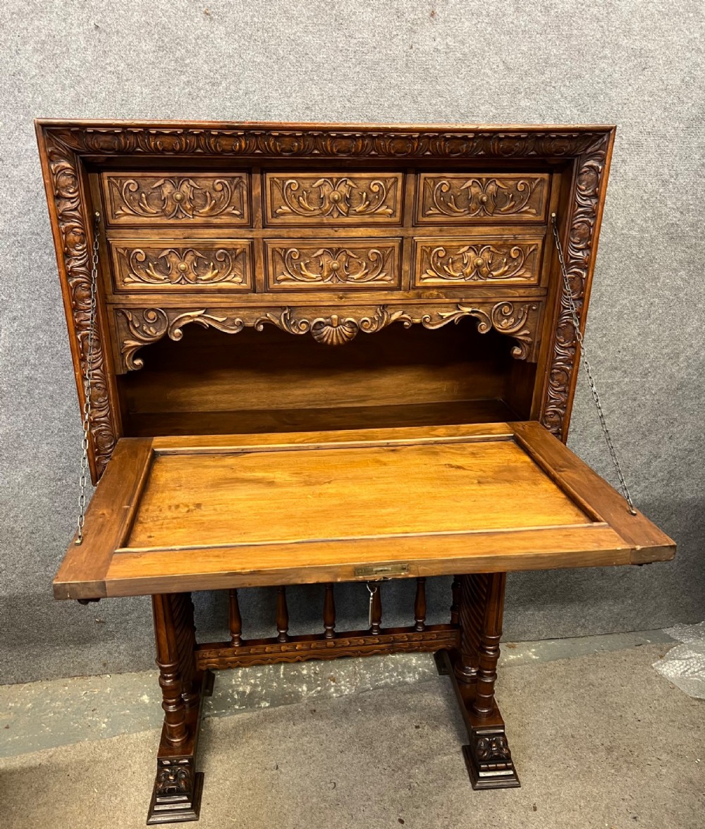 A carved Spanish vargueno cabinet in dark wood, featuring relief figures on the drop-front flap, decorative brass hardware, and a turned trestle stand. View IV