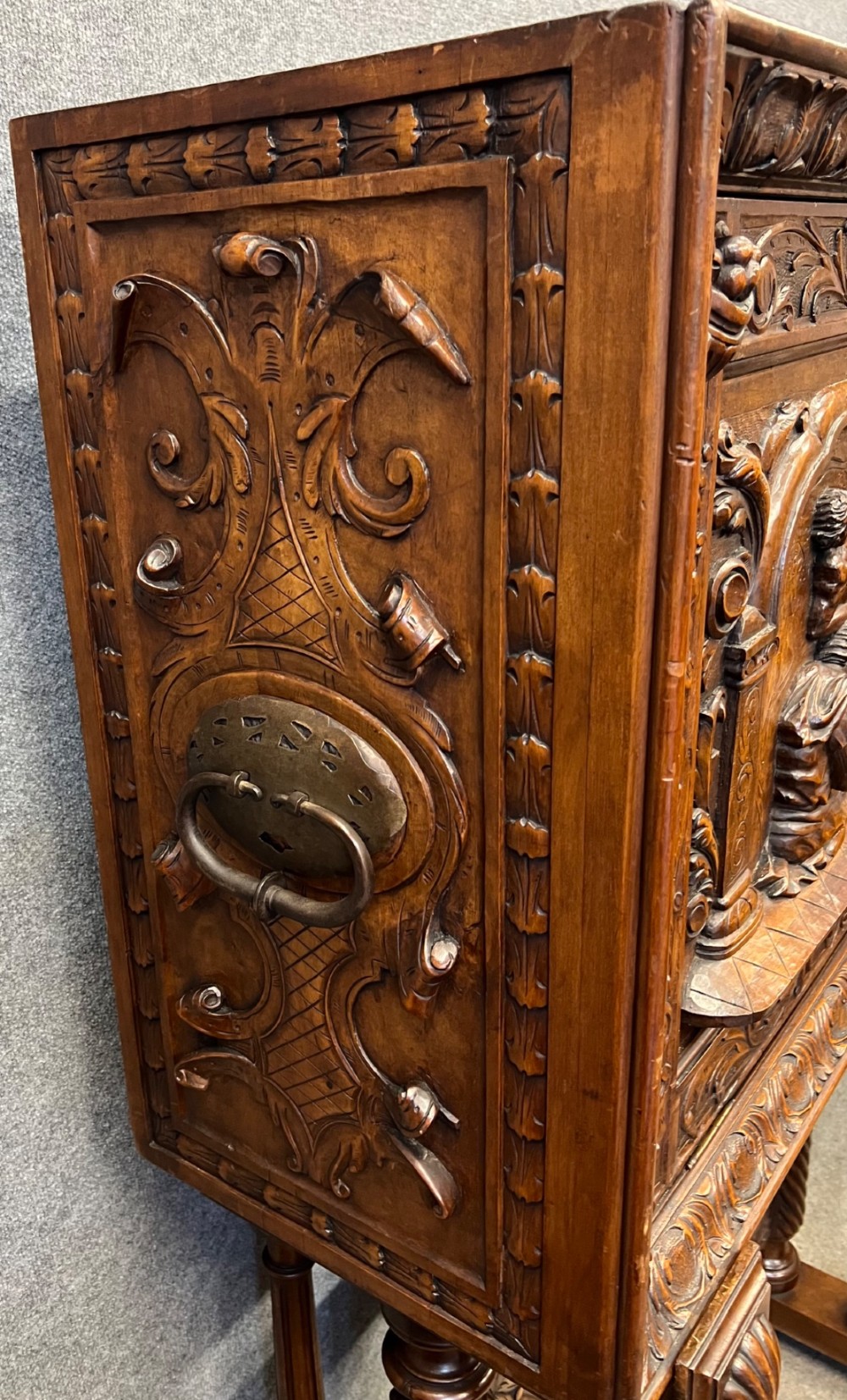 A carved Spanish vargueno cabinet in dark wood, featuring relief figures on the drop-front flap, decorative brass hardware, and a turned trestle stand. View IX