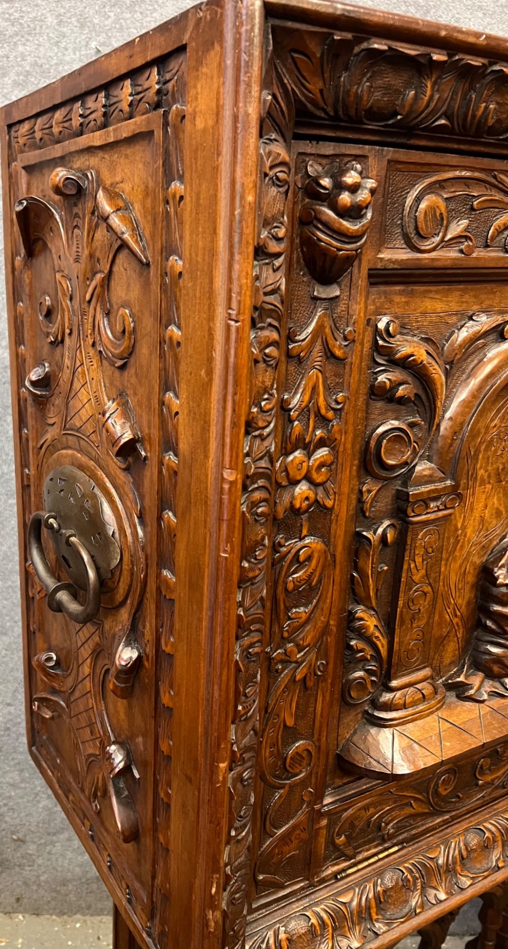 A carved Spanish vargueno cabinet in dark wood, featuring relief figures on the drop-front flap, decorative brass hardware, and a turned trestle stand. View XII