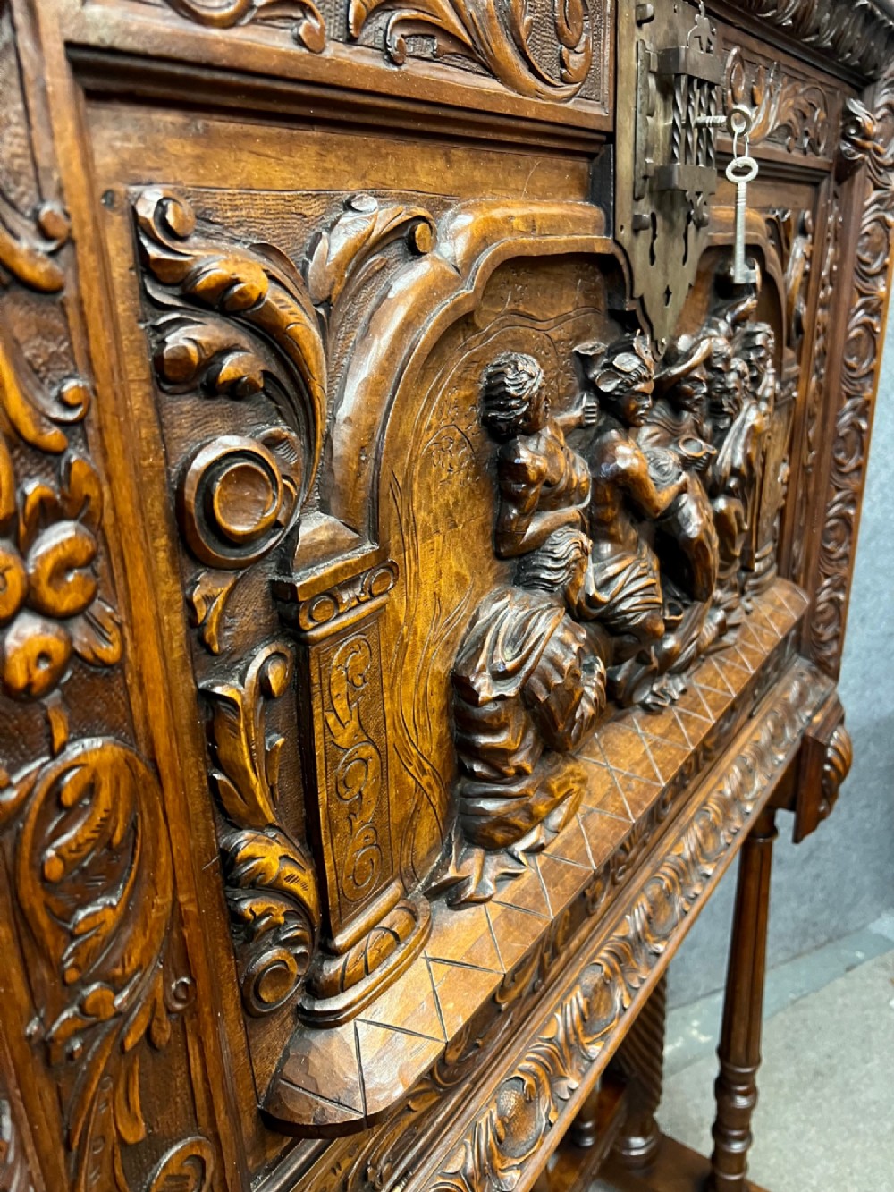 A carved Spanish vargueno cabinet in dark wood, featuring relief figures on the drop-front flap, decorative brass hardware, and a turned trestle stand. View XIII