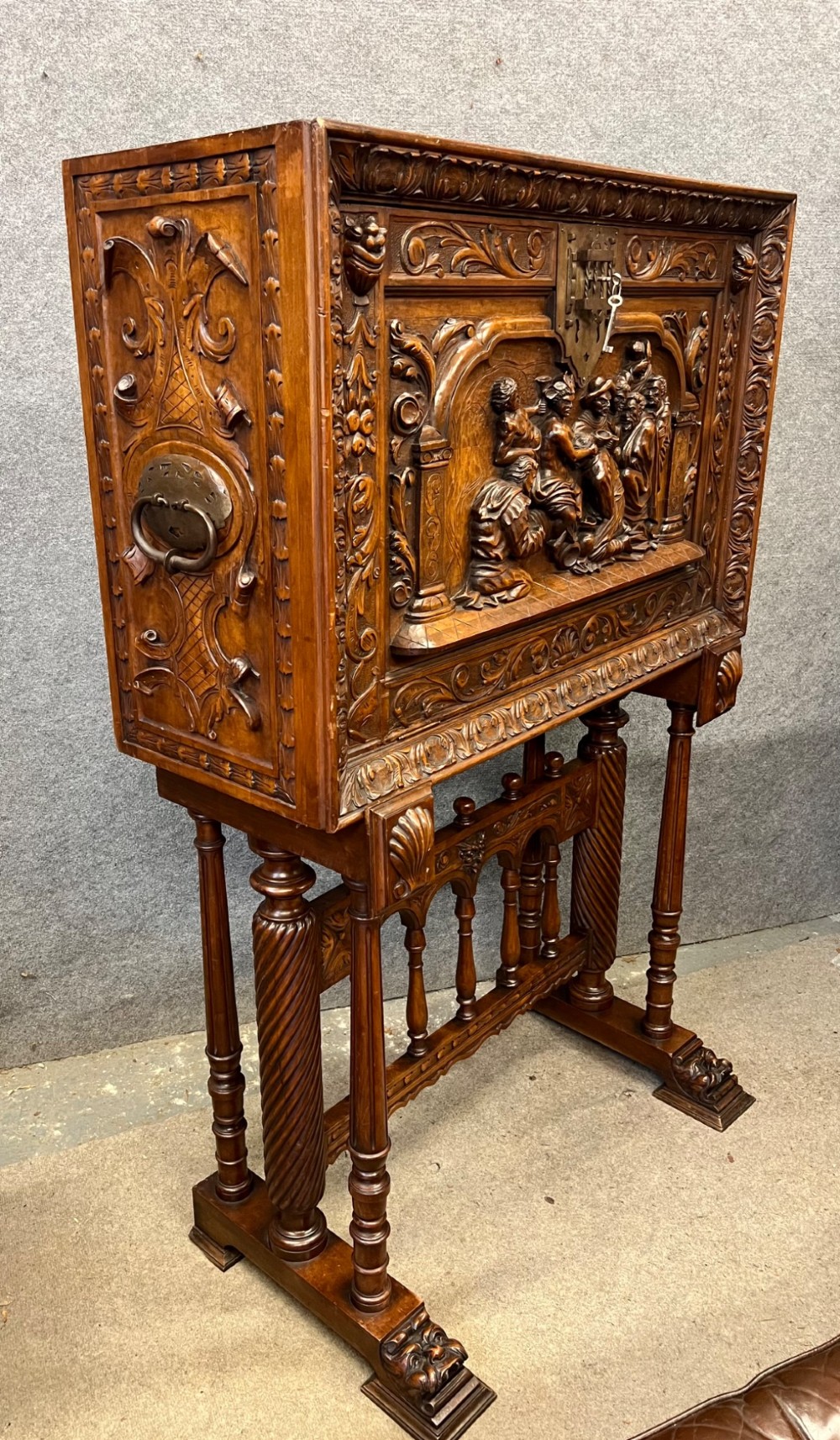 A carved Spanish vargueno cabinet in dark wood, featuring relief figures on the drop-front flap, decorative brass hardware, and a turned trestle stand. View XIV
