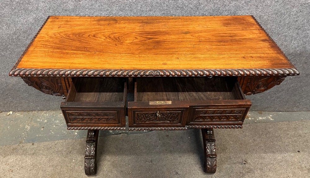 An Anglo-Indian carved hardwood centre table featuring a polished rectangular top, rope-twist carved borders, relief-carved frieze drawers, and detailed trestle legs. View IV