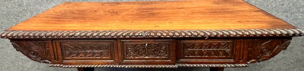 An Anglo-Indian carved hardwood centre table featuring a polished rectangular top, rope-twist carved borders, relief-carved frieze drawers, and detailed trestle legs. View VIII