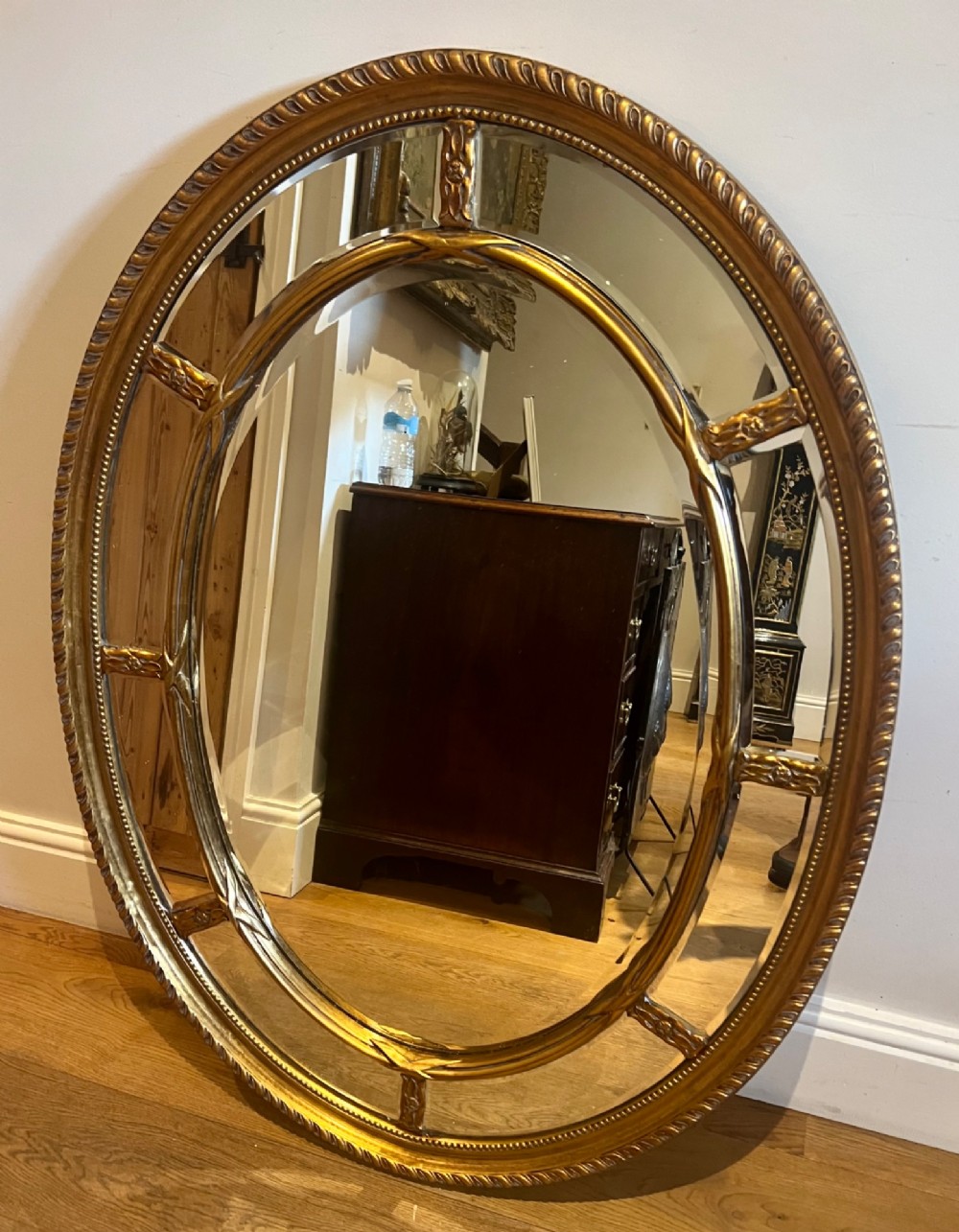 An oval gilded wall mirror featuring a bevelled central mirror surrounded by outer glass margin panels and a gadrooned rim.