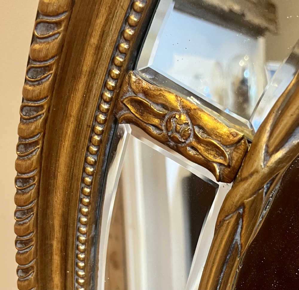 An oval gilded wall mirror featuring a bevelled central mirror surrounded by outer glass margin panels and a gadrooned rim. View II