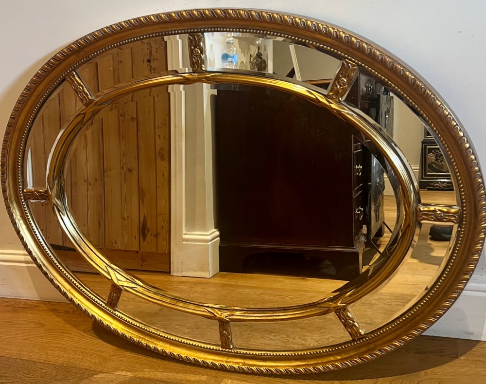 An oval gilded wall mirror featuring a bevelled central mirror surrounded by outer glass margin panels and a gadrooned rim. View VII