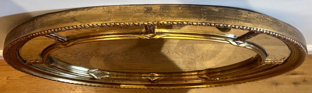 An oval gilded wall mirror featuring a bevelled central mirror surrounded by outer glass margin panels and a gadrooned rim. View VIII