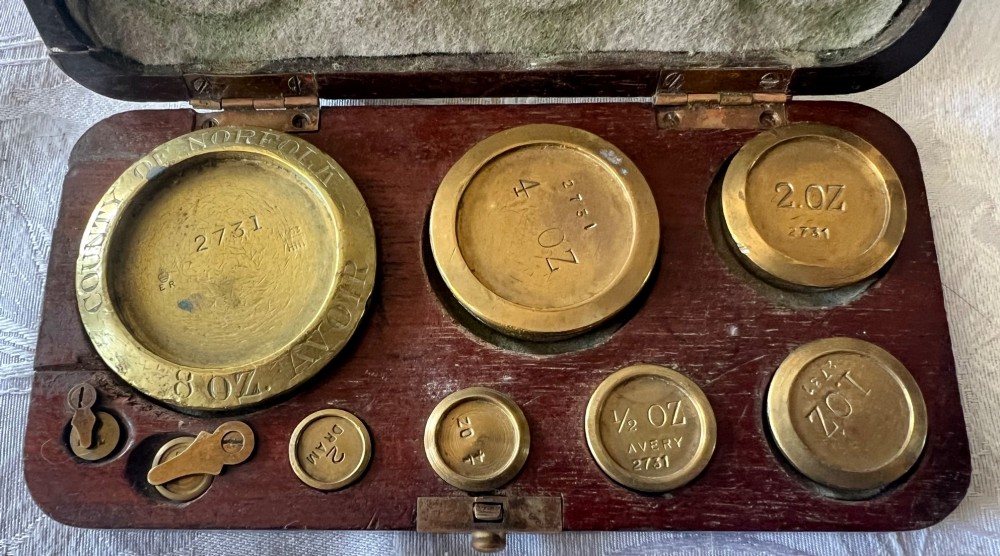 An open Edwardian wooden box lined with green baize, housing stamped brass imperial weights. View V