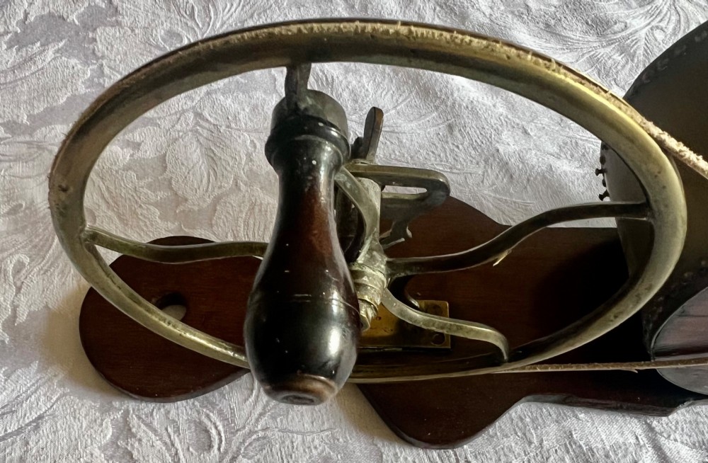 An antique mechanical rotary fireplace bellows featuring a mahogany wooden base, a brass spoked wheel with a hand crank, a driving band, and a brass casing leading to a tapered nozzle. View V