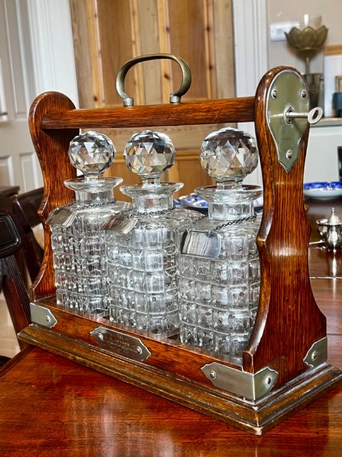 EDWARDIAN OAK TANTALUS WITH THREE GLASS DECANTERS MATCHING HALLMARKED SILVER LABELS