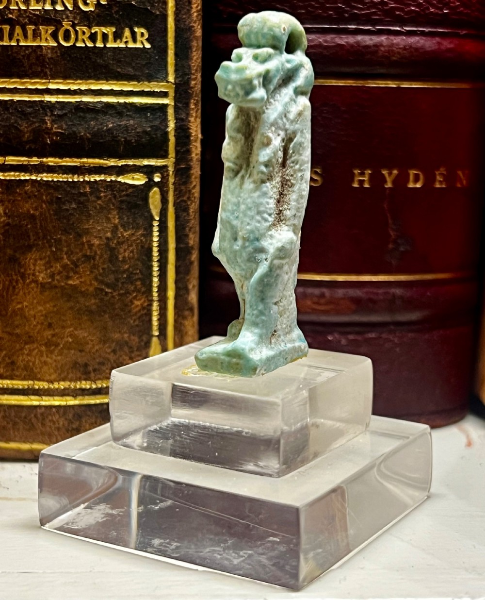 A small, turquoise-glazed ancient Egyptian amulet mounted upright on a clear acrylic stand against a blurred book background.