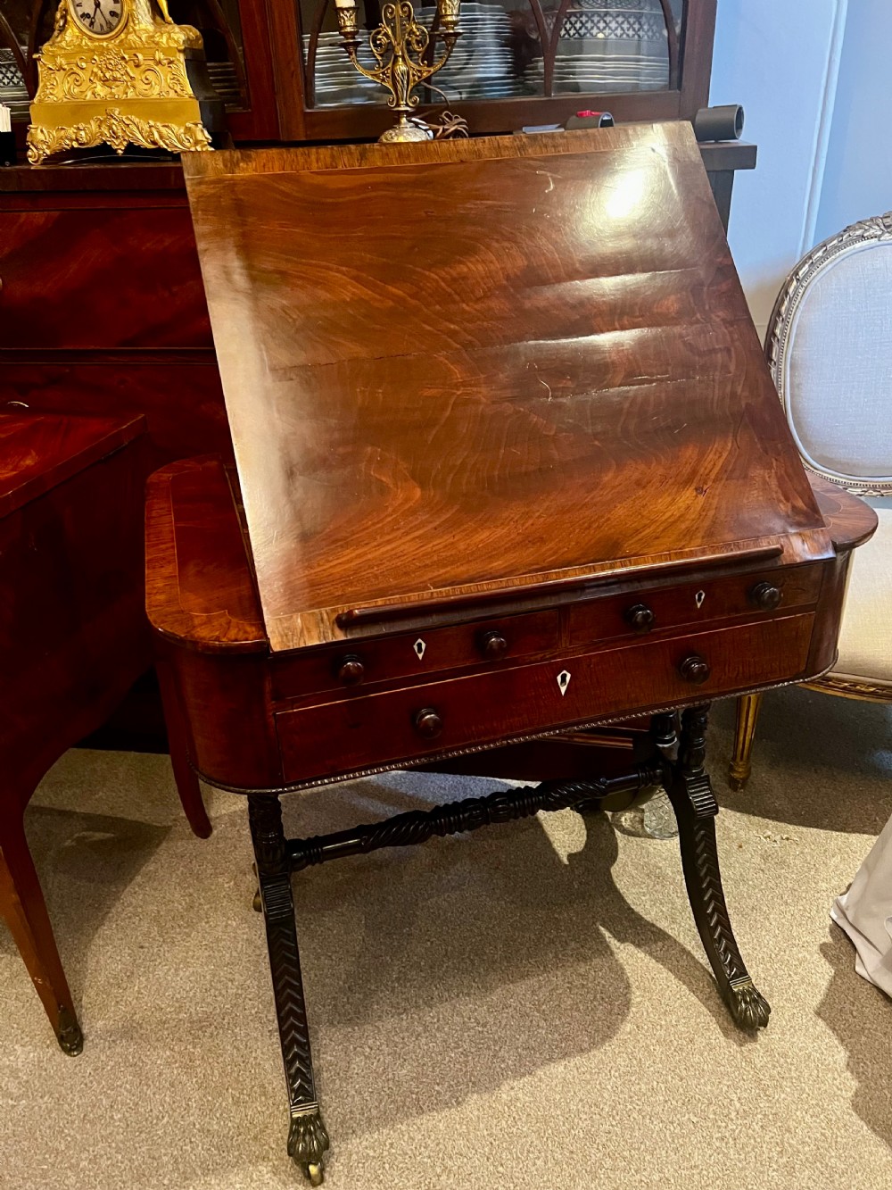 An antique dark wood Regency reading table featuring an adjustable sloped top, small drawers with brass key escutcheons, and carved intersecting legs with castors.