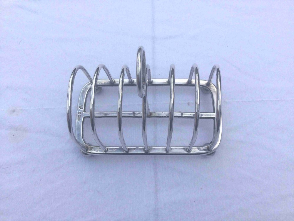 A silver six-slot toast rack featuring arched wire dividers, a central oval loop handle, and four small ball feet resting on a plain background. View V