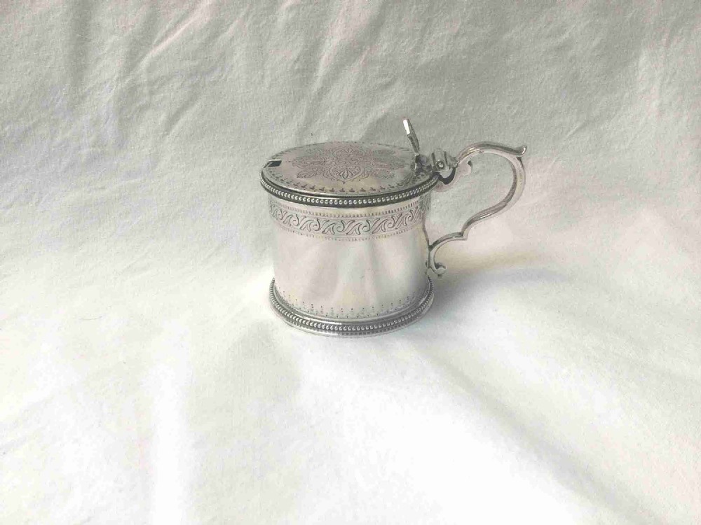 A cylindrical sterling silver mustard pot featuring bright-cut engraved patterns on the flat lid, beaded rims, and a scrolled handle.