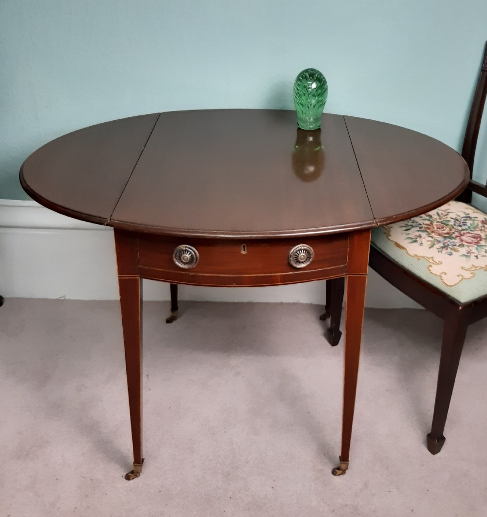 An 18th-century Georgian oval mahogany Pembroke table featuring two drop leaves, a single drawer with round brass handles, tapered legs, and brass castors.