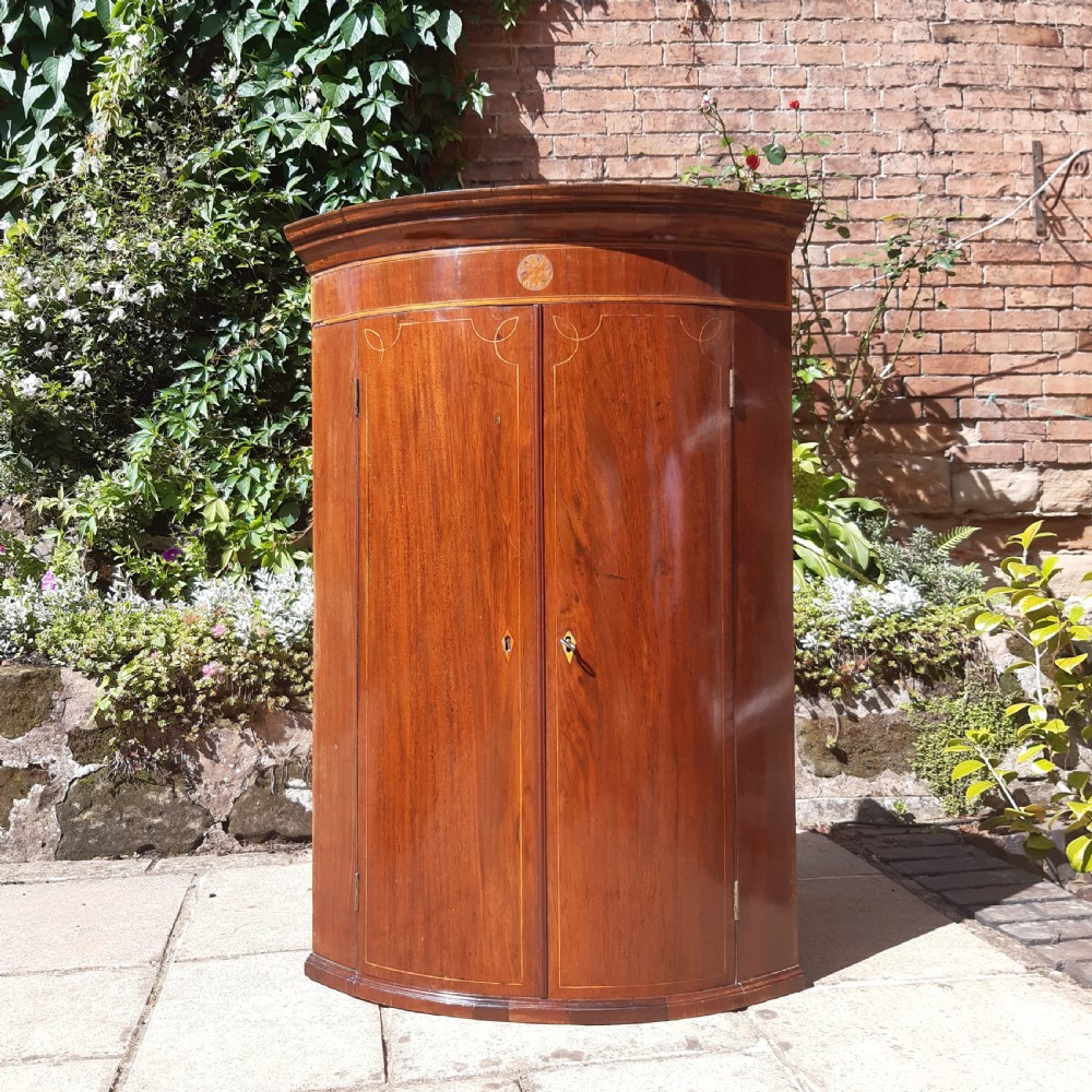 An antique Georgian mahogany corner cupboard featuring a curved bow-front facade, inlaid double doors, and brass escutcheons.