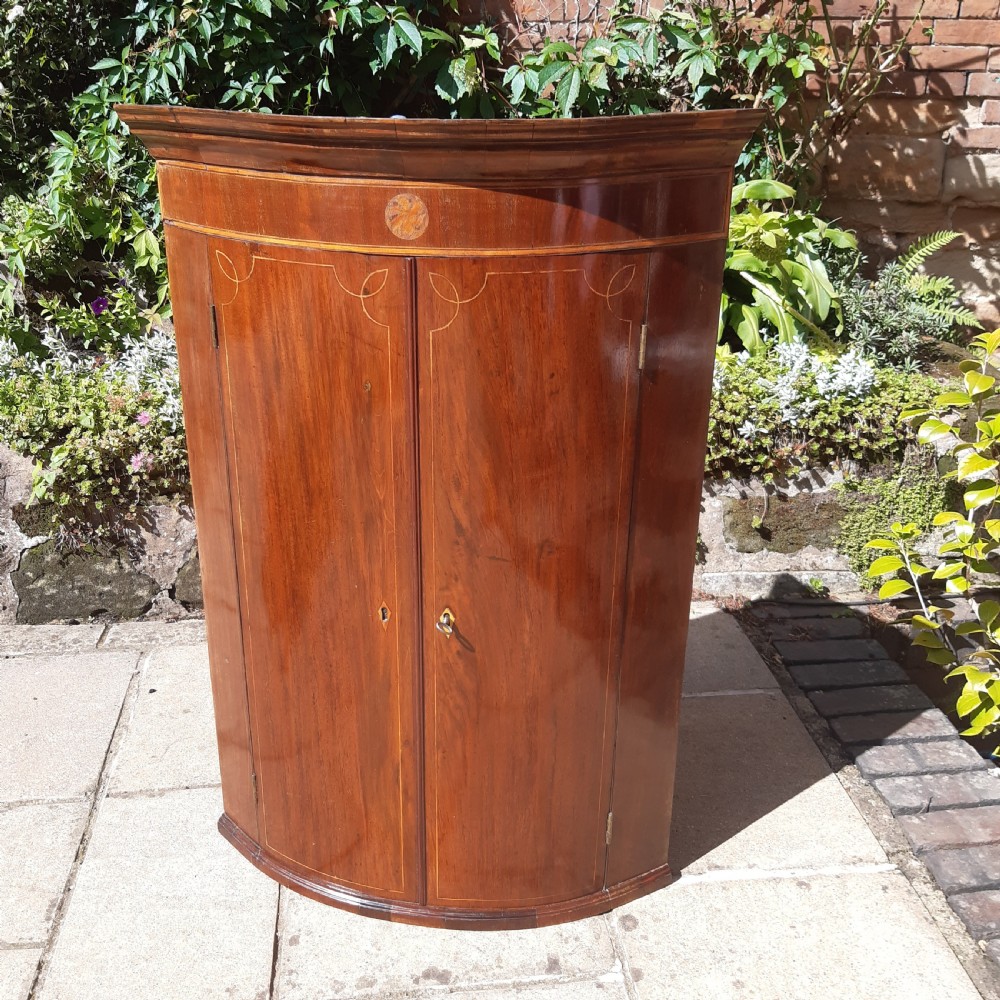 An antique Georgian mahogany corner cupboard featuring a curved bow-front facade, inlaid double doors, and brass escutcheons. View II