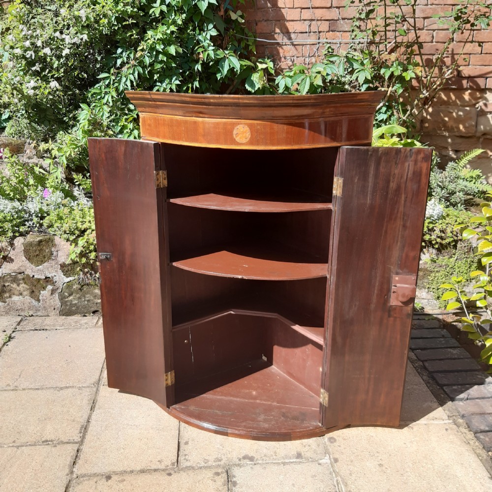 An antique Georgian mahogany corner cupboard featuring a curved bow-front facade, inlaid double doors, and brass escutcheons. View III