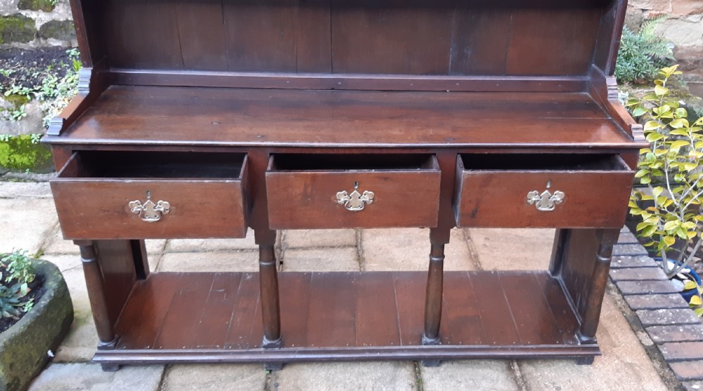 A dark oak antique dresser featuring an open plate rack with three shelves above three frieze drawers, supported by turned pillars over an open potboard base. View III