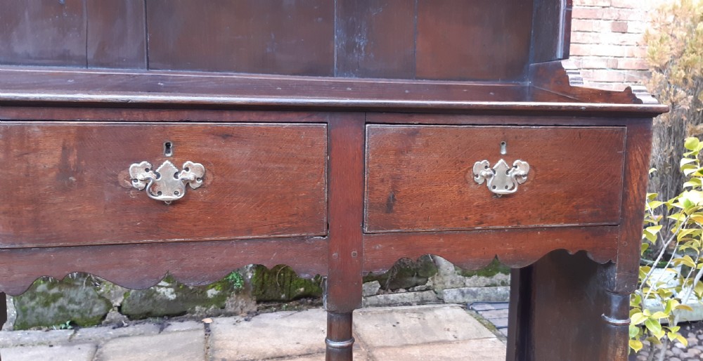A dark oak antique dresser featuring an open plate rack with three shelves above three frieze drawers, supported by turned pillars over an open potboard base. View IV