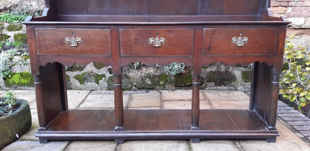 A dark oak antique dresser featuring an open plate rack with three shelves above three frieze drawers, supported by turned pillars over an open potboard base. View V