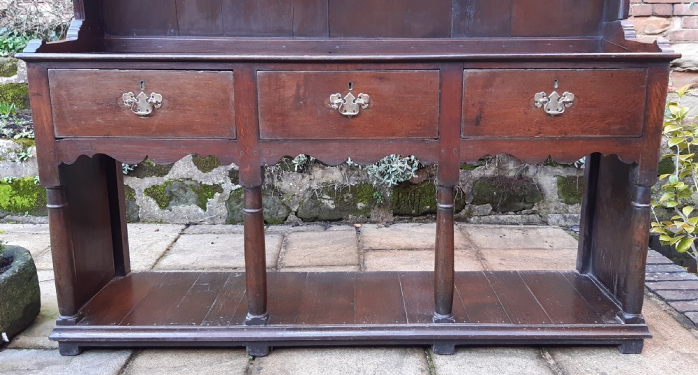 A dark oak antique dresser featuring an open plate rack with three shelves above three frieze drawers, supported by turned pillars over an open potboard base. View XV