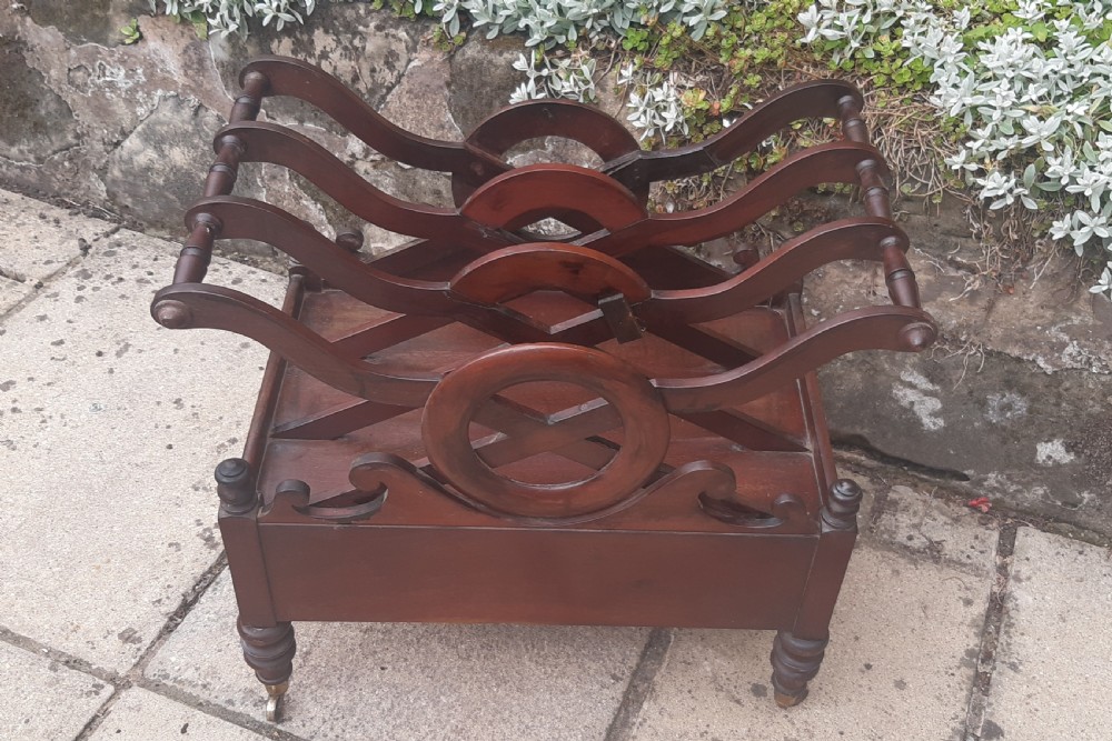An early nineteenth-century mahogany Canterbury with curved slatted compartments, a central carved circular motif, a single drawer, and turned legs on brass castors. View II