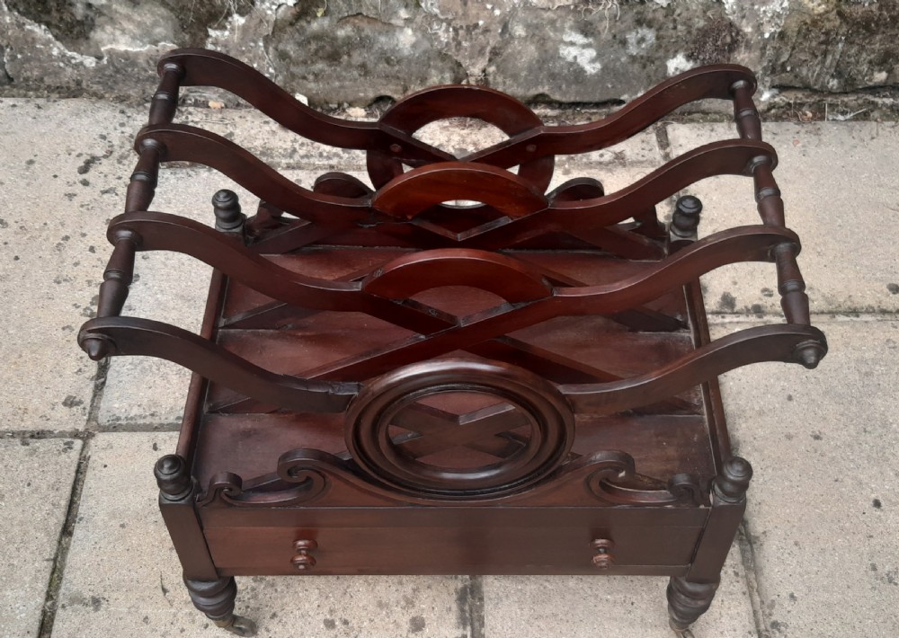 An early nineteenth-century mahogany Canterbury with curved slatted compartments, a central carved circular motif, a single drawer, and turned legs on brass castors. View V