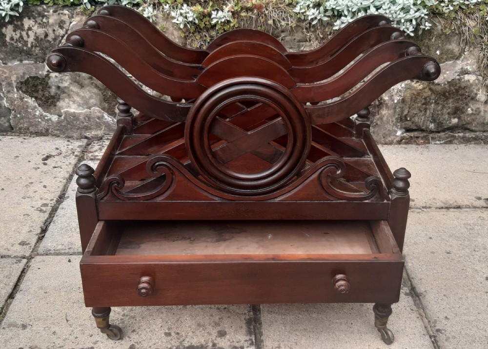 An early nineteenth-century mahogany Canterbury with curved slatted compartments, a central carved circular motif, a single drawer, and turned legs on brass castors. View VII