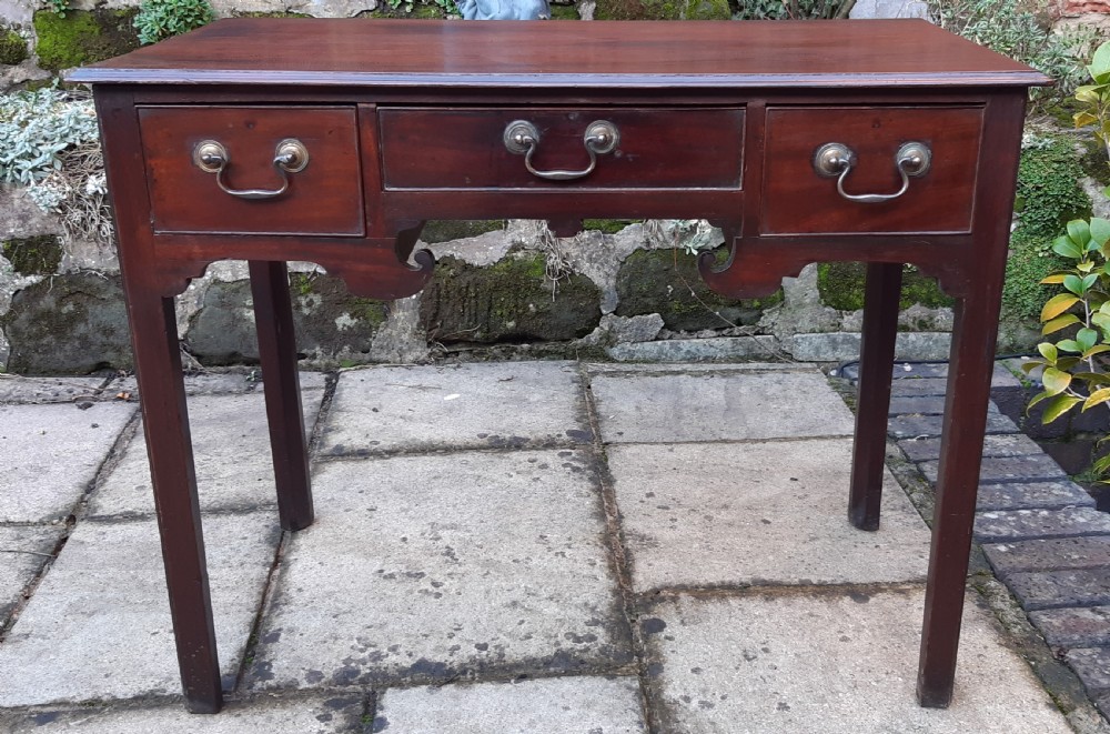 An antique Georgian dark mahogany lowboy featuring a rectangular top, three drawers with brass swan-neck handles, a shaped apron, and straight square legs.