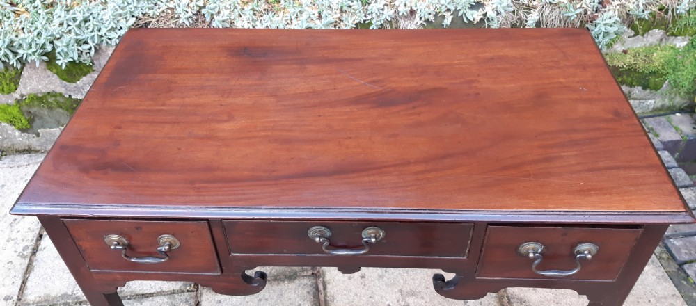 An antique Georgian dark mahogany lowboy featuring a rectangular top, three drawers with brass swan-neck handles, a shaped apron, and straight square legs. View V