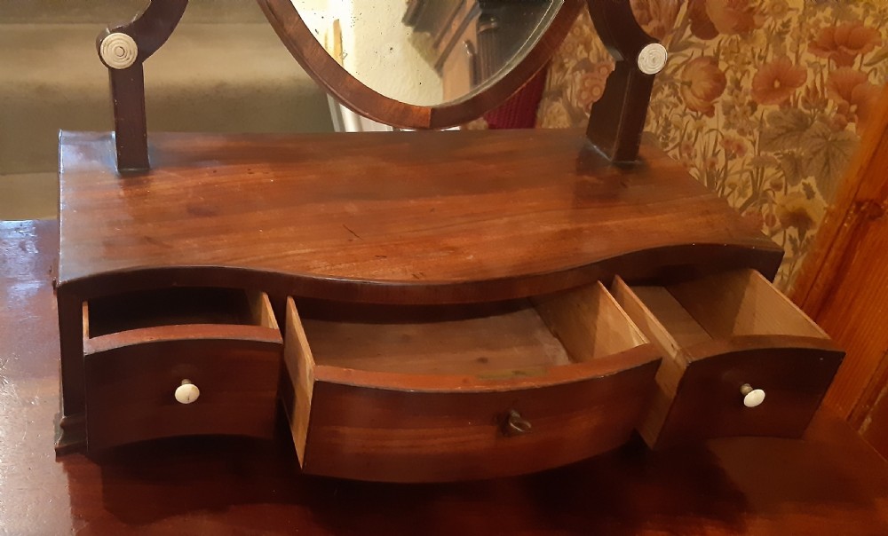 An antique mahogany toilet mirror featuring a shield-shaped swivelling frame set on curved uprights above a serpentine-front base with small drawers. View II