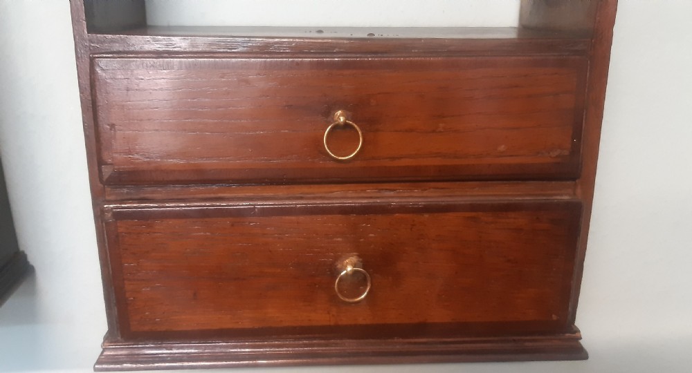 A dark oak wall-mounted shelf unit featuring two open tiers with decorative pierced corner brackets above two small drawers fitted with brass ring handles. View V