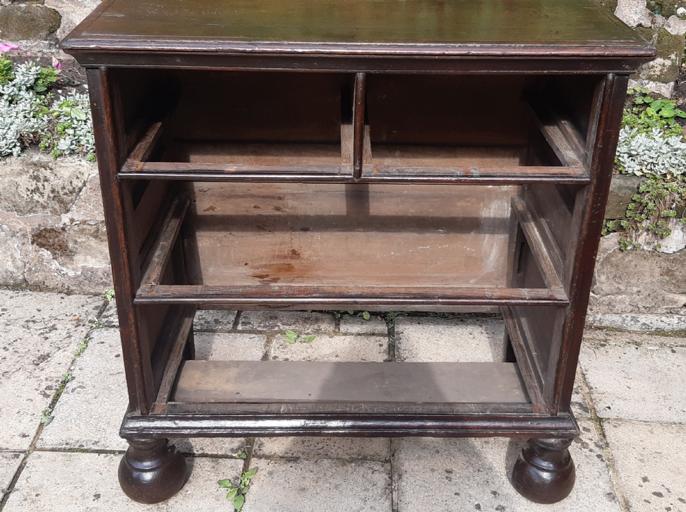 A 17th-century dark oak chest of drawers featuring geometric panelled drawer fronts, brass drop handles, keyhole escutcheons, and turned bun feet. View IV