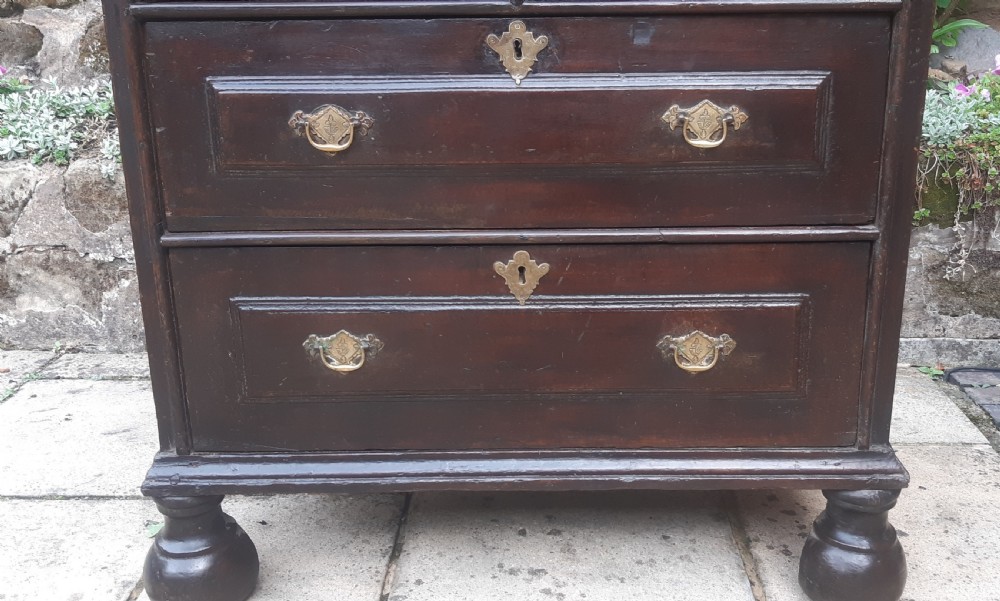 A 17th-century dark oak chest of drawers featuring geometric panelled drawer fronts, brass drop handles, keyhole escutcheons, and turned bun feet. View V