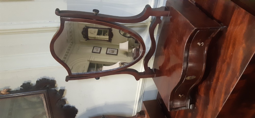 An antique mahogany toilet mirror featuring an adjustable swivel glass frame held by curved uprights above a serpentine drawer base with brass hardware. View II