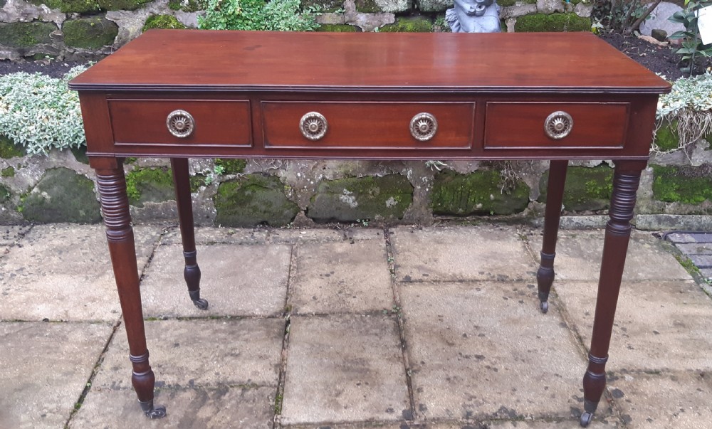 A Regency mahogany writing table featuring three shallow frieze drawers with brass rosette handles, supported by reeded turned legs on brass castors.
