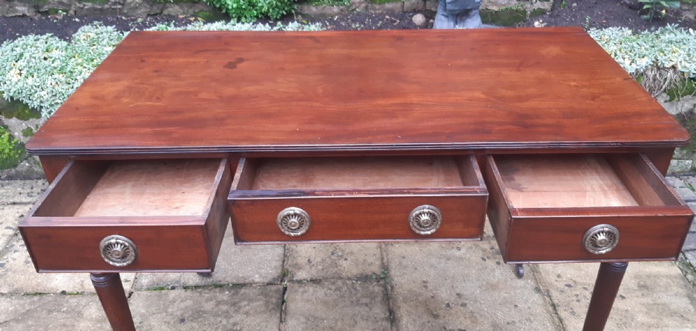 A Regency mahogany writing table featuring three shallow frieze drawers with brass rosette handles, supported by reeded turned legs on brass castors. View II