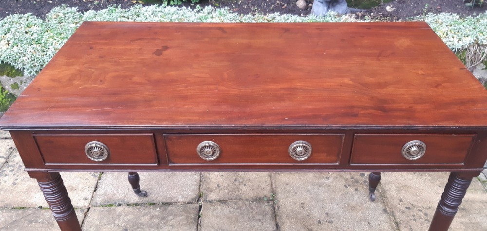 A Regency mahogany writing table featuring three shallow frieze drawers with brass rosette handles, supported by reeded turned legs on brass castors. View IV