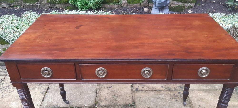 A Regency mahogany writing table featuring three shallow frieze drawers with brass rosette handles, supported by reeded turned legs on brass castors. View VI