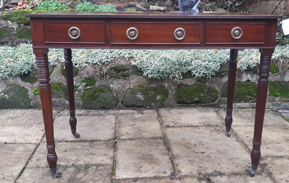 A Regency mahogany writing table featuring three shallow frieze drawers with brass rosette handles, supported by reeded turned legs on brass castors. View VII