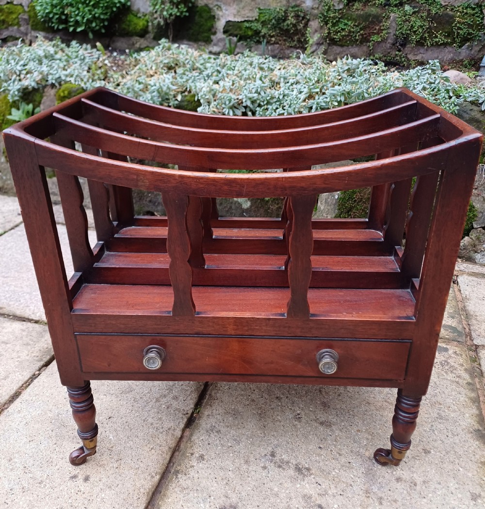A Regency mahogany music canterbury featuring curved slatted divisions, a base drawer with two brass knobs, turned legs, and brass castors.