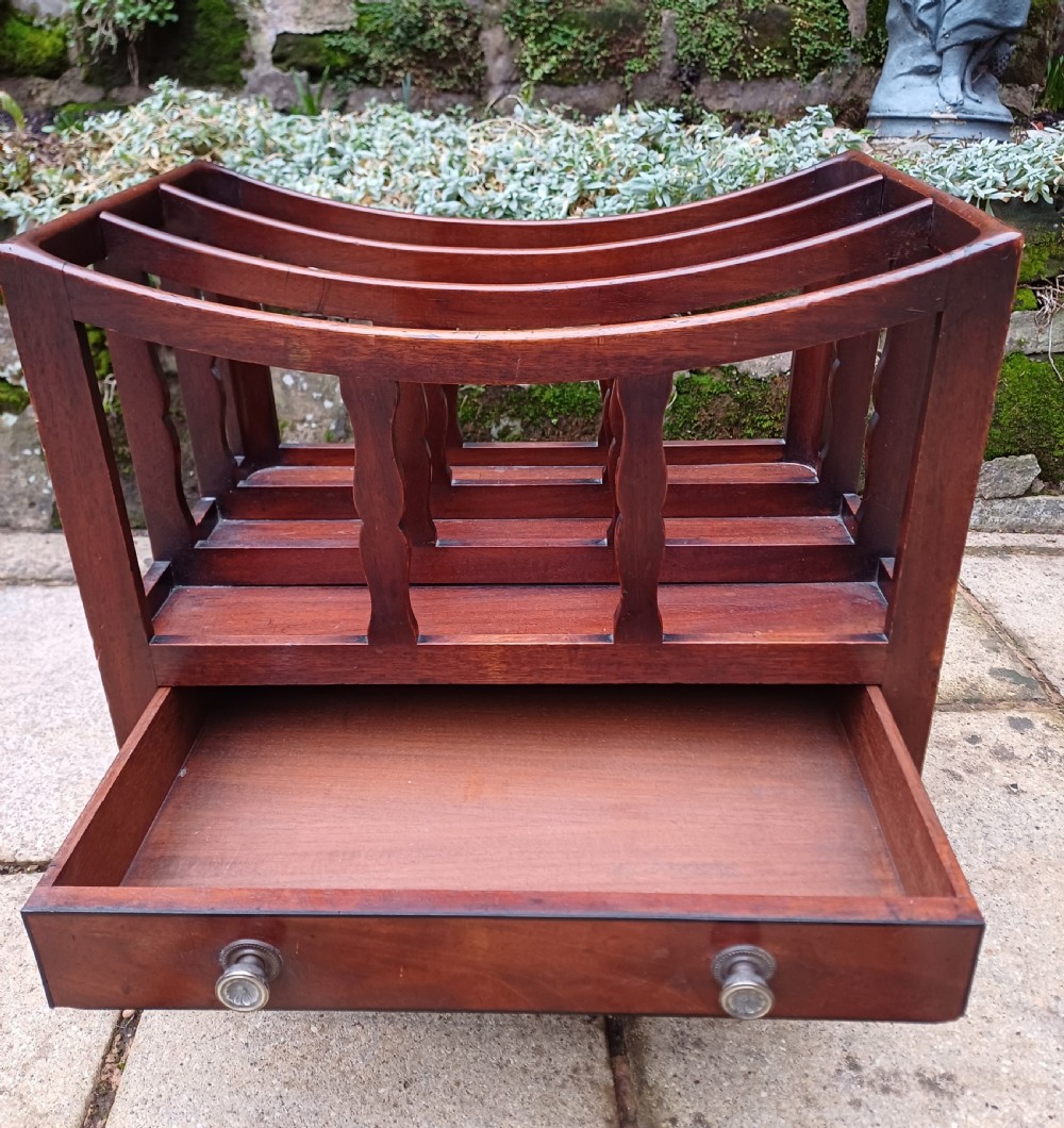 A Regency mahogany music canterbury featuring curved slatted divisions, a base drawer with two brass knobs, turned legs, and brass castors. View V