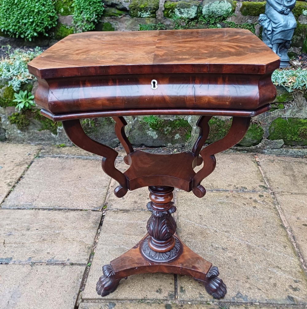 A Victorian flame mahogany work table featuring a shaped top with a central keyhole escutcheon, supported by C-scroll arms, a carved acanthus pedestal, and a triform base with claw feet.